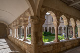 <center>Le cloître de Saint-Genis des Fontaines.</center>Le cloître roman se compose de quatre piliers d'angle et de quatre piliers médians reliés par des arcatures portant sur colonnes isolées. Piliers et colonnes s'établissent sur un mur bahut. Les arcs sont surmontés de constructions postérieures. Le tout est en marbre. Les chapiteaux sont ornés de figures grossières.
