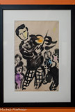 <center>Le Violoneux.</center>1957. Encre de Chine et crayon gras sur papier Achat, 2023.
Le Violoneux accroît la collection du musée sur la période vençoise de Chagall (1950-1965), riche d'œuvres bibliques, mais aussi l'ensemble des œuvres qui illustre le thème musical à l'instar des nombreux personnages musiciens tels le Roi David et sa harpe, les anges musiciens, ou encore les violonistes.
Chagall est installé à Vence depuis 1950. En 1957, il réalise le dessin Le Violoneux, qui désigne un violoniste de village. Ce personnage habite les œuvres de l'artiste depuis sa jeunesse, lui rappelant les fêtes de son enfance au shtetl. A cette période, il travaille sur le cycle du Message Biblique (1956-1966), mais poursuit parallèlement ses recherches sur les thèmes du cirque et de la musique, qui ont toujours été liés dans ses œuvres. La peinture murale pour le foyer du Théâtre de Francfort, intitulée Commedia dell'arte, inaugurée en 1959, met en lumière l'univers musical du cirque, tout comme La Danse et Le Cirque bleu, réalisées en 1950, pour un projet pour le Watergate Theater de Londres. La musique représente pour Chagall un art original qui révèle la vie dans son énergie la plus pure. C'est ce qui compose l'essence même du Violoneux : un violoniste joue une musique endiablée au milieu de personnages qui l'écoutent avec attention. Chagall fait la part belle à la ligne noire, contrastée, qui est relevée de discrètes touches colorées.
