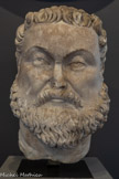 <center>Musée Saint-Raymond</center>Tête quasi colossale de Maximien Hercule (?)
Vers 250-310
Empereur de 286 à 305
Marbre de Saint-Béat (Haute-Garonne) après 293. <br>
En 293, Dioclétien instaure un mode de gouvernement nouveau : la Tétrarchie (pouvoir à quatre têtes). Maximien, Auguste d'Occident, était épaulé par un César, Constance Chlore, et Dioclétien, Auguste d'Orient, par un autre César, Galère. L'ensemble de la décoration présentée dans cet espace, ainsi que ces quatre portraits, dépendent certainement de cette période et témoigneraient d'une restauration fastueuse de la villa de Chiragan sous Maximien, dit Hercule. Sur ce portrait, les mèches de la frange, des tempes et de la barbe rappellent la tête de l'Hercule combattant Diomède du grand cycle sculpté exposé ici-même. La statue dont dépendait cette tête devait atteindre 2,75 m de hauteur ; elle était donc plus grande que la seconde représentation de cet empereur dans la villa : un relief, restitué au centre de cet espace
