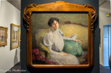 <center>Portrait de madame Alice Tissier</center>HENRI MARTIN (1860-1943). Huile sur toile, 1900.
Le genre du portrait n’intéressa Henri Martin que lorsque les modèles étaient ses proches : sa femme, ses enfants ou quelques amis. Ici le modèle est une amie de la famille, jeune femme brune an sombre regard, Alice Tissier pose, assise, de trois quarts race, sur un canapé Louis XV, en une attitude sans doute familière. Avec une attention évidente, Henri Martin individualise son visage éclairé par une lumière vive venant de droite; le jeu ombre/lumière «ovalise » le visage et met l'accent sur un nez droit un peu fort. Elle nous regarde attentive et rêveuse à la fois, tête à peine inclinée, sourire esquissé. La ligne serpentine et souple des moulures du bois du sofa, légèrement de biais par rapport au mur brun et neutre à l'arrière-plan, forme une sorte d'écrin ouvert sur la droite. Le vert vif du coussin sur lequel repose son bras droit, l’étoffe blanche et fine d'une robe très iooo aux manches gigot, les fleurs, aux pétales mousseux rose grenat, que l’on aperçoit au premier plan à gauche et à droite, animent et réchauffent l’harmonie brune et jaune ocre du reste de la toile, par ailleurs assez terne. Ici la pose et le décor pourtant très sobre adoucissent avec bonheur le caractère d'un visage sans doute en réalité très charpenté.