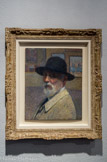 <center>Autoportrait</center>HENRI MARTIN, (1860-1943). Huile sur panneau.
Henri Martin qui ne fut pas particulièrement intéressé par le genre du portrait, contre toute attente, se portraitura toute sa vie. On compte une dizaine de ces exercices. L’Autoportrait présenté date de la dernière décade de sa vie. Ce tableau surprend par son originalité : ce n'est pas un portrait mais une présence, une sorte d'instantané photographique. Dans un premier temps en effet, c'est la silhouette d'un vieil homme coiffe d'un chapeau cachant en partie son visage qui s'impose. C'est une sorte d'anti-portraiture de la littéralité des traits ou de la force psychologique d'un regard, ici absent.
Contrairement à d'autres autoportraits, où le peintre pose dans sa fonction peignant une toile dans son atelier, ici, il se présente en « monsieur », dans le cadre d'un Salon ou d'une galerie. Sur le mur, à l'arrière-plan en effet sont accrochés des tableaux; les siens sans doute. Peut-être est-ce la façon pour Henri Martin de suggérer une notoriété assumée.