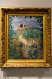 <center>Nu à la fontaine</center>LEBASQUE (1865-1937). Huile sur toile, 1911
Henri Lebasque n'ignore rien de l’avant-garde de son temps. Comme les « poulains » du marchand Ambroise Vollard (1866-1939), il fait dès 1906 des essais de céramique chez André Metthey (1871-1920) à Asnières où il décore des vases et des plats.
D'abord paysagiste, Lebasque est particulièrement connu pour son travail sur les nus et sur la couleur. Lebasque regroupe ici deux thèmes qui lui sont chers dans ce que le critique d'art Adolphe Tabarant (1863-1950) appelle un « paysage animé » qui n'est ni véritablement un nu, et pas davantage un paysage. Comme il le remarque justement, Lebasque renouvelle le thème de la jeune fille gracile. Ici nue et de profil, les bras relevés, la figure occupe la place centrale de la composition. Elle se recoiffe, la tête penchée. Son bras dissimule son visage, totalement absent. Son chapeau repose à ses côtés. A l'arrière-plan, on distingue dans des camaïeux de bleus quelques nuages au-dessus de montagnes qui surplombent un plan d'eau tranquille. La scène semble totalement irréelle. Seule une silhouette de femme portant une robe rose sur le chemin de terre ancre la composition dans la réalité.