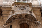 <center>Villefranche-de-Rouergue </center>Maison Gaubert. Au-dessus de la porte, un arc est décoré de feuilles d'acanthe et de calots sculptés. Des animaux y sont représentés dans une corbeille végétale. L'un porte un harnais (un cheval), un second tient dans sa gueule une branche de pommier qui pourrait évoquer le péché originel. Derrière eux sont figurés un personnage et un dragon. La scène, qui semble s'inspirer de l'histoire religieuse, illustrerait une fable populaire représentant des animaux savants.