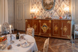 <center>La Salle à manger </center>Grand buffet à cinq vantaux avec placage de bois de violette, marbre Rance et bronze doré. La façade est tripartite, à ressaut plat central. Elle est liée aux côtés rectangulaires par des pans coupés droits sur plan curviligne. Le dessus de marbre épouse la découpe générale. Le placage de bois consiste, sur la façade, en des filets et chevrons autour d'un large méplat central (délimité par une moulure de bronze) à marqueterie en damier coupé de diagonales (les intersections en bois plus foncé), sur les pans coupés, en une marqueterie en damier coupé de diagonales, sur les côtés, en des filets et du frisage pour le panneau central. Le décor de bronzes ciselés et dorés est composé de filets marquant les quatre pieds d'angle, d'une forte moulure courant autour du piètement du meuble reprise en un filet à la ceinture sous le marbre, et sur la façade de huit écoinçons ajourés à angle droit et crosse.