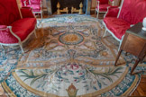 <center>Chambre sur cour.</center>Tapis comportant un caisson central carré à rentrées concaves aux quatre axes, en moulure or, à centre d'acanthes claires alternées de ruban rose en torsade. Le masque central est auréolé d'or, dans une couronne de lauriers vert foncé. Autour sont disposées quatre coquilles prolongées d'une torche or à flammes entre des guirlandes roses à feuillage foncé. À chaque extrémité prend place une lyre à bras en acanthe et base en marbre rose, sommé d'une coquille en or avec une couronne de lauriers et un ruban bleu, posée sur deux arcs et deux carquois à flèches entre des guirlandes roses et vertes foncées. Aux deux axes sont placées deux palmes à ruban rouge et contours de lauriers à fleurs entre deux moulures or et acanthes à chaque angle. La bordure est manquante. Ce tapis, symbolisant Apollon, est la partie centrale d'un tapis plus grand qui devait prendre place dans la Grande Galerie du Louvre. Il fait partie d’un ensemble de quatre-vingt-treize tapis de neuf mètres de long commandé à Le Brun par Colbert. Les cartons furent réalisés aux Gobelins notamment par François Francart et Baudrin Yvart. Pour produire cet ensemble colossal, dont le tissage dura vingt ans (1670-1689), le ministre de Louis XIV réorganisa la manufacture de la Savonnerie, en réunissant à Chaillot deux ateliers existants, celui des Dupont au Louvre, et celui des Lourdet.