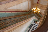<center>L'escalier d'honneur.</center>Lustre à douze bras de lumière suspendu au plafond au moyen d'un fût orné de branches de laurier retombant sur le socle, servant de soutien à trois figures d’enfants traités en ronde-bosse, adossés au fût. Ceux-ci soutiennent à bout de bras, dans un geste d’effort renforcé par une jambe repliée contre le fût, des chaînes censées compléter l’armature. Les bras de lumière sont disposés en deux registres concentriques et affectent une légère différence de hauteur, permettant d’augmenter l’impression de profusion qui caractérise ce lustre dont les boules placées sur les tronçons des fûts cannelés servant de bobèches, rappellent les sphères lisses des globes. Le traitement sophistiqué de l’ensemble, que termine une pomme de pin entourée de feuilles à la base du lustre, est surtout dû au dessin très fin des rinceaux formant les bras. 1850-1874.